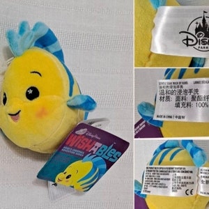 May include: Yellow and blue plush toy of a fish with a smiling face and sparkly eyes. The toy is labeled "Disney Parks" and "Wishables".