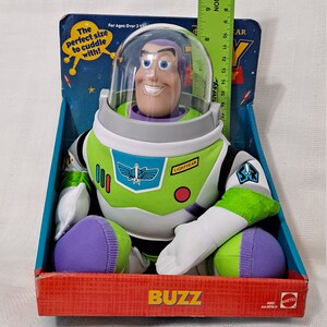 K&ouml;nnte beinhalten: Ein Pl&uuml;sch-Buzz Lightyear Spielzeug aus der Toy Story-Reihe. Das Spielzeug sitzt in einer Schachtel mit den Worten "Buzz" und "Lightyear" darauf. Das Spielzeug tr&auml;gt einen gr&uuml;n-wei&szlig;en Raumanzug mit einem violetten Helm. Das Spielzeug ist f&uuml;r Kinder ab 3 Jahren geeignet.
