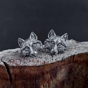May include: A pair of silver-toned fox head stud earrings. The earrings are detailed with textured fur and facial features. The earrings are displayed on a piece of weathered wood against a dark background.