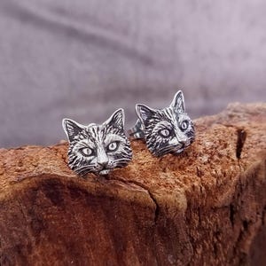 Op de afbeelding: Een paar zilverkleurige kattenkop-oorstekers. De oorbellen zijn gedetailleerd met geëtst haar en gezichtskenmerken, waaronder ogen, neus en snorharen. De oorbellen worden gepresenteerd op een stuk hout met een natuurlijk, getextureerd oppervlak.