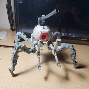 Może przedstawiać: Biały i szary robot z czerwonym okiem i długą szarą bronią. Robot ma sześć nóg i stoi na drewnianej powierzchni.