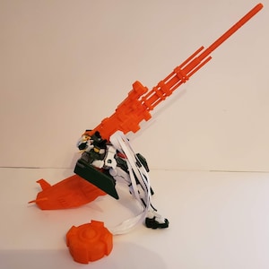 以下が含まれることがあります： 大きなオレンジ色の武器が付いた、詳細なロボットのモデル。ロボットは緑、白、黒のアクセントがあります。ベースや円形の部品など、追加のオレンジ色のコンポーネントがロボットの近くに配置されています。