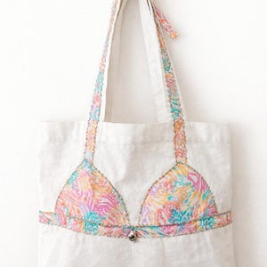 Op de afbeelding: Witte tote bag met een kleurrijk bikinitopontwerp. De bikinitop heeft een levendig patroon van roze, oranje en blauwgroen. De bandjes en handvatten zijn versierd met dezelfde stof met patroon, en een kleine strik siert de handvatten.