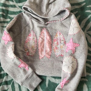 May include: A gray hooded sweatshirt with pink and white fabric appliqués. The front features three surfboard shapes with floral and lace details. The sleeves have starfish and sand dollar appliqués. The hood and seams have pink stitching.