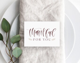 Puede incluir: Un montaje de mesa con un plato blanco, una servilleta de lino y una tarjeta con la frase "Thankful For You" en cursiva. Cuchara y cuchillo de color cobre. Una ramita de eucalipto y una bellota añaden un toque natural.