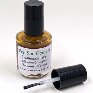 Peut inclure: Une bouteille de Pen Sac Cement avec un bouchon noir et un applicateur à pinceau. La bouteille contient un liquide brun doré et porte une étiquette avec du texte. Le bouchon est retiré, révélant le pinceau.