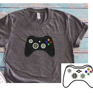 May include: A grey t-shirt with a black and white embroidered video game controller. The controller has four buttons, a directional pad, and two analogue sticks.