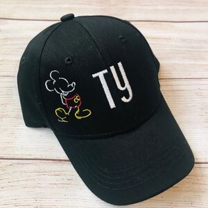 Op de afbeelding: Zwarte baseballpet met een witte geborduurde Mickey Mouse en de naam "TY" in wit.