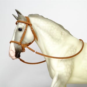 Puede incluir: Imagen detallada de una figura de caballo gris claro con una brida de cuero marrón. La brida tiene anillos metálicos plateados y se ajusta alrededor de la cabeza del caballo. El caballo tiene una nariz rosada y ojos oscuros.