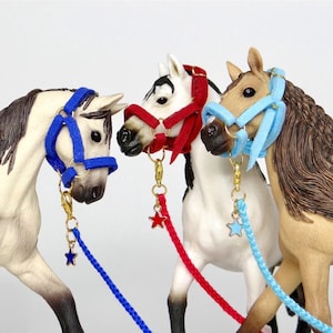May include: Three toy horses with colourful halters and leads. The horses are painted in shades of brown, white, and black. Each horse has a different coloured halter: blue, red, and light blue. The leads are also colour-coordinated with star-shaped charms.