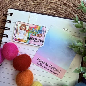 May include: A colorful library card sticker with a cartoon illustration of a person holding an apple. A magnetic bookmark with the text "Magnetic Bookmark" and a barcode are also visible. The image is on a notebook with colorful felt balls.