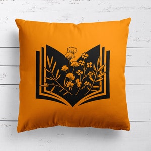 May include: Orange throw pillow with a black silhouette of an open book with flowers growing out of it.