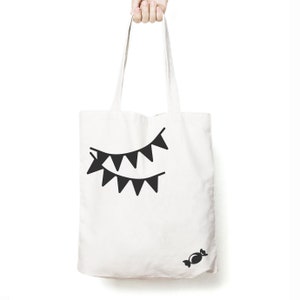 May include: A white canvas tote bag featuring a black graphic print of two rows of triangular bunting flags positioned in the upper centre and a small black graphic of a wrapped sweet located in the lower right corner of the bag.