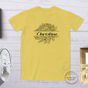 以下が含まれることがあります： 黄色のTシャツに、黒い輪郭の菊の花と「Christine」という名前がプリントされています。