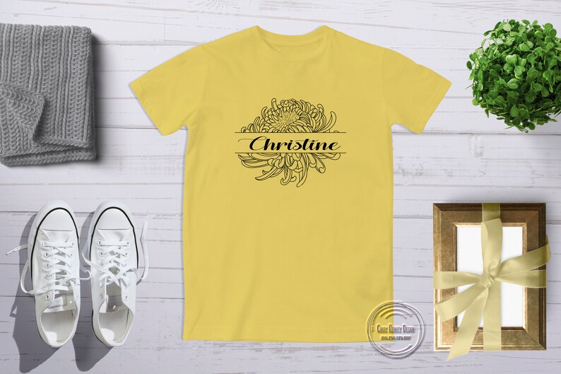 以下が含まれることがあります： 黄色のTシャツに、黒い輪郭の菊の花と「Christine」という名前がプリントされています。