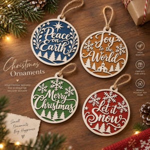 Op de afbeelding: Vier ronde houten kersthangers met jute touwtjes. Het blauwe ornament toont Peace on Earth, het goudkleurige Joy to the World, het groene Merry Christmas en het rode Let it Snow, allemaal voorzien van uitgesneden dennenbomen en sneeuwvlokken.