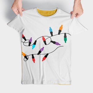 Puede incluir: Camiseta blanca con un dise&ntilde;o de luces navide&ntilde;as de colores enhebradas.