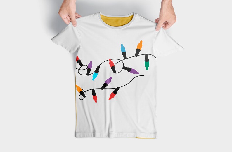 Puede incluir: Camiseta blanca con un dise&ntilde;o de luces navide&ntilde;as de colores enhebradas.
