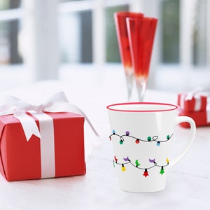 Puede incluir: Una taza de cer&aacute;mica blanca con un borde rojo y un dise&ntilde;o de luces navide&ntilde;as de colores. La taza est&aacute; sobre una mesa blanca junto a una caja de regalo roja con una cinta blanca.