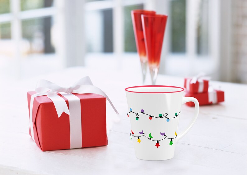 Puede incluir: Una taza de cer&aacute;mica blanca con un borde rojo y un dise&ntilde;o de luces navide&ntilde;as de colores. La taza est&aacute; sobre una mesa blanca junto a una caja de regalo roja con una cinta blanca.
