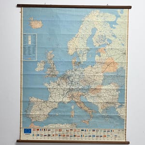 May include: Vintage map of Europe, showing countries and road networks, with a light blue background. The map is mounted on wooden rods. Includes a list of flags and a legend. The map is designed for hanging.