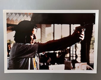 May include: A black and white photograph of a person holding a gun. The person is wearing a white shirt and has long dark hair. The background is blurry.