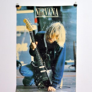 Puede incluir: Un póster de la banda Nirvana. La imagen muestra a una persona con pelo largo y rubio sosteniendo una guitarra eléctrica negra. El nombre de la banda está impreso en letras mayúsculas blancas en la parte superior del póster.