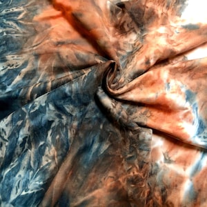 Puede incluir: Primer plano de una tela con un patrón de tie-dye en tonos azules, marrones y naranjas. La tela está doblada y arrugada, creando un aspecto texturizado.
