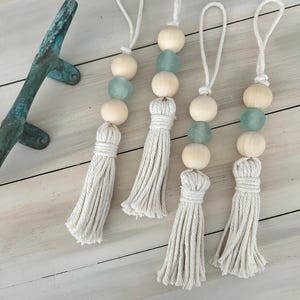 May include: Four decorative tassels with wooden beads and sea glass accents. Each tassel is made of cream-colored rope and features a loop for hanging. The beads are light wood and the sea glass is a pale blue-green color.