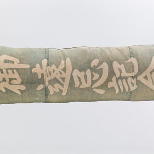 May include: A long, rectangular pillow made of a light blue fabric with white Japanese characters printed on it. The pillow is made of a woven fabric with a slight texture.