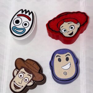 May include: Four character-themed shoe charms featuring a white spork with red and blue details, a red-haired character, a brown cowboy hat-wearing character, and a purple-hooded character with a white face, all with a glittery finish.