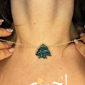 Puede incluir: Collar de cadena dorada con un colgante de árbol verde. El colgante es una representación detallada de un cedro. El collar se lleva alrededor del cuello. La palabra "لجين" está escrita en árabe.