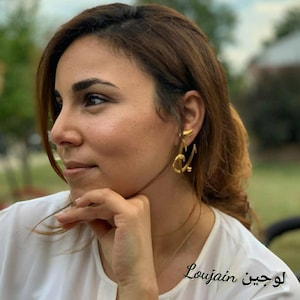 Peut inclure: Boucles d'oreilles dorées en forme des lettres "L" et "J" portées à l'oreille. Les boucles d'oreilles font partie d'une collection de bijoux. Le nom "Loujain" est écrit en arabe au bas de l'image.