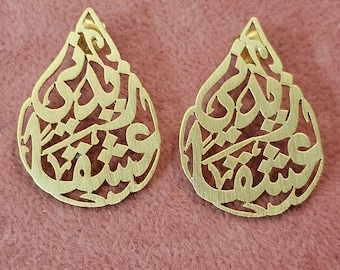 May include: Two gold-colored teardrop-shaped earrings. Each earring features intricate Arabic calligraphy cut-out designs. The earrings are displayed on a pink surface.