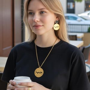 Peut inclure: Une personne portant un t-shirt noir et des bijoux dorés, dont un collier et des boucles d'oreilles avec un pendentif circulaire portant l'inscription arabe. La personne porte également un bracelet et une bague dorés, et tient une tasse de café blanche.