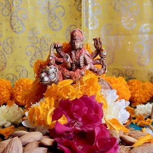 May include: A brass statue of a deity with multiple arms, atop a lion, surrounded by orange and yellow flowers. A bowl of almonds and a red rose are in the foreground. The background is a yellow fabric with a silver paisley pattern.