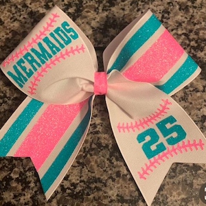 May include: White cheer bow with pink and teal stripes. The bow has the words "MERMAIDS" and the number "25" on it, both in teal.