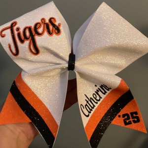 Cheerleading bow designed in custom colors and text. Bow features team name "Tigers" on upper loop. Name on tail stripe with year on triangle. Orange and Black personalized bow for Gameday, Varsity Cheer or Senior Night.