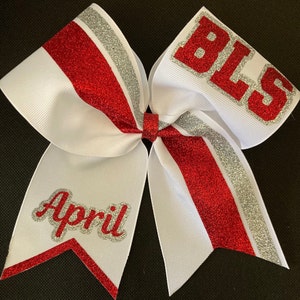 May include: White cheerleading bow with red and silver glitter accents. The bow features the text "BLS" in red glitter on one side and the name "April" in red glitter on the other side. The bow is made of a white ribbon.