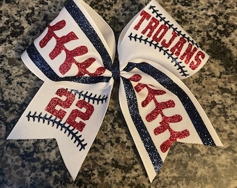May include: White cheer bow with red and blue glitter. The bow has a baseball design with the words "Trojans" and the number "22" on it.