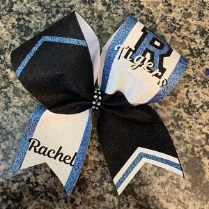 May include: A black and white cheer bow with blue glitter accents. The bow has the text "TR Tigers" on one side and "Rachel" on the other side.