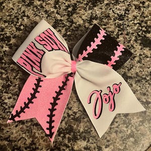 May include: A large, decorative hair bow with a baseball theme. The bow features white and pink ribbons with the text "PINK SOX" and "JoJo" in pink. The bow has black stitching details.