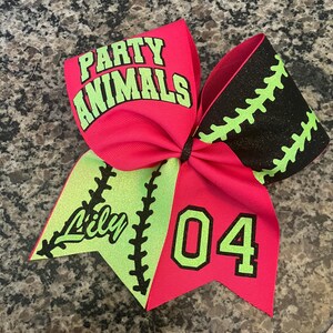 May include: A vibrant pink and black cheer bow with the words "PARTY ANIMALS" in green. The bow features the name "Lily" and the number "04" in green. The bow has a baseball stitch design.