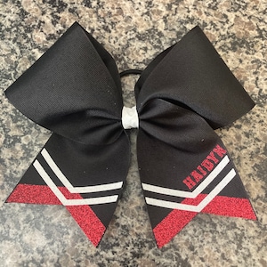 May include: Black cheer bow with white stripes and red glitter accents. The bow has the name "Hadyn" written on it.