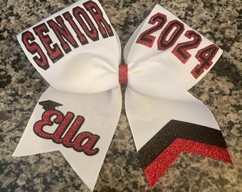 May include: White cheer bow with red and black glitter accents. The bow has the text "Senior 2024" on one side and "Ella" with a graduation cap on the other side.