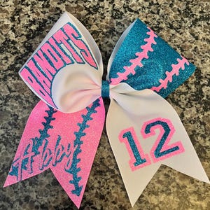 May include: A white cheer bow with pink and teal glitter. One side has the word "Bandits" in teal glitter on a white background. The other side has the number "12" in teal glitter on a white background. Both sides have a teal and pink baseball stitch design.