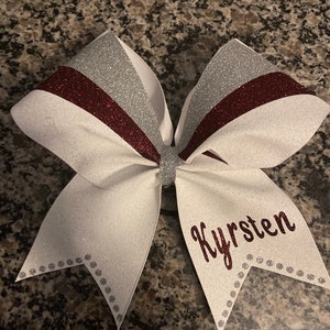 May include: White cheer bow with silver and burgundy stripes and silver glitter. The bow has the name "Kyirsten" written in burgundy glitter.