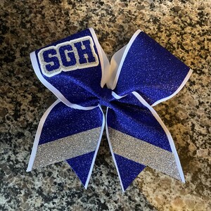 May include: A blue and silver glitter cheer bow with a white center and the letters "SGH" in white on a silver background.