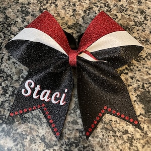 May include: A black cheer bow with red and white stripes and red glitter accents. The bow has the name "Staci" embroidered in white on the front.