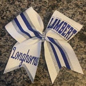 May include: White cheer bow with silver and blue glitter stripes. The bow has the words "Lambert Longhorns" in blue glitter lettering.
